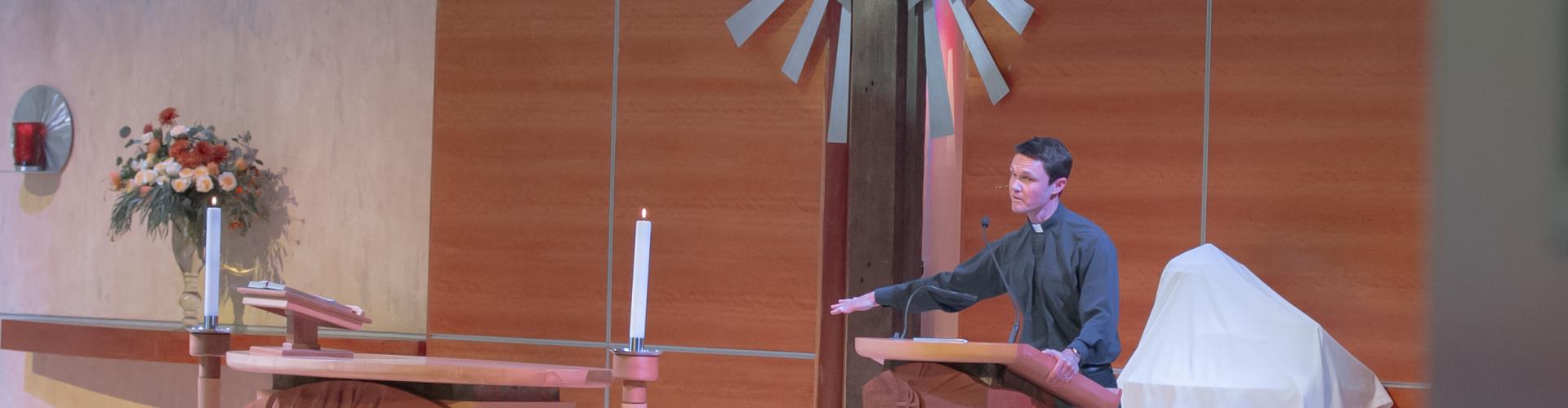 Pastor Garrett Siemsen stands at pulpit looking at congregation while preaching. A large wooden cross is to his right.