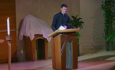 Pastor Garrett wears a dark shirt with clerical collar and a dark jacket. He leans on ambo while delivering sermon