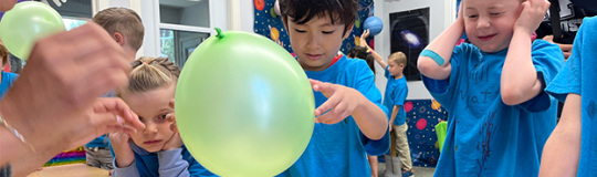 child holds balloon while another child has his hands over his ears and his eyes tightly shut in anticipation of balloon breaking